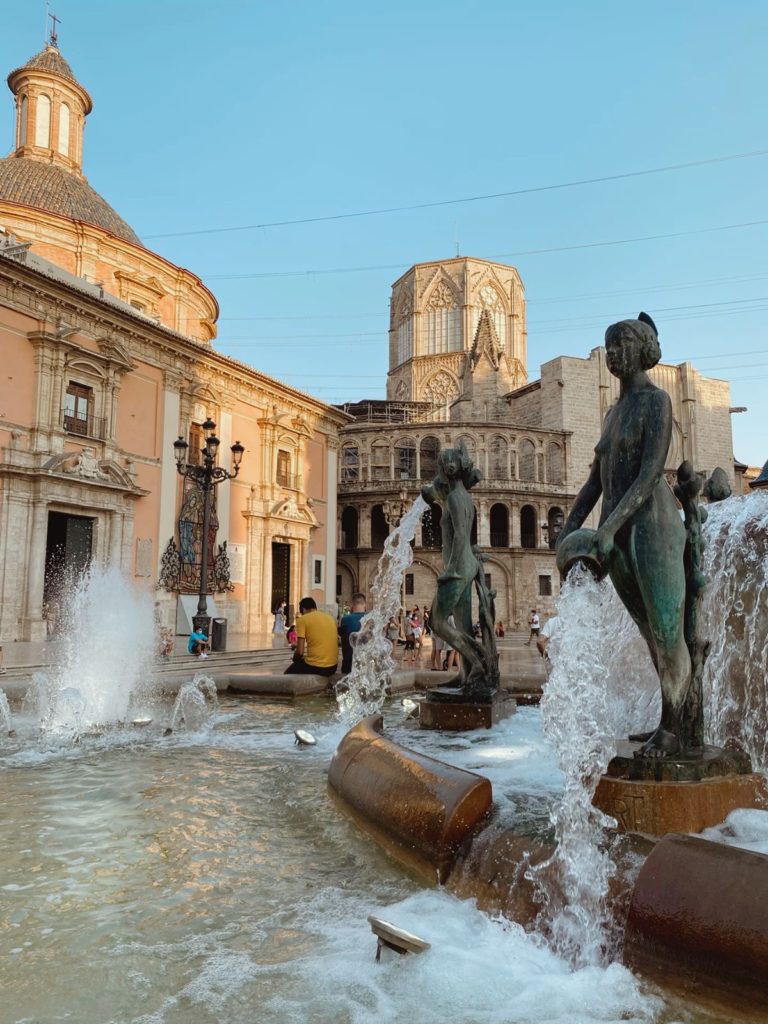 Fontaine du Turia au premier plan avec ses statues de bronze déversant de l'eau, située sur la Plaza de la Virgen à Valence, Espagne. En arrière-plan, la Basilique de la Virgen de los Desamparados et la cathédrale de Valence sous un ciel bleu clair.