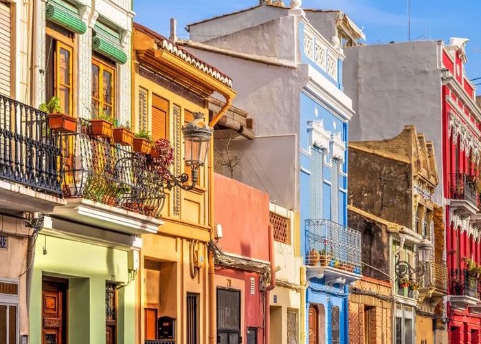 Une rangée de maisons de ville étroites et mitoyennes aux façades vivement colorées (vert pistache, jaune ocre, bleu ciel et rouge vif) sous un ciel bleu dégagé. Les bâtiments présentent des balcons en fer forgé ornés de plantes en pots, des volets traditionnels et des lampadaires muraux de style ancien.